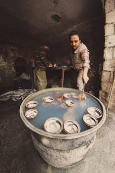 A man cleans metal dishes in a rustic workshop setting, showcasing traditional techniques.