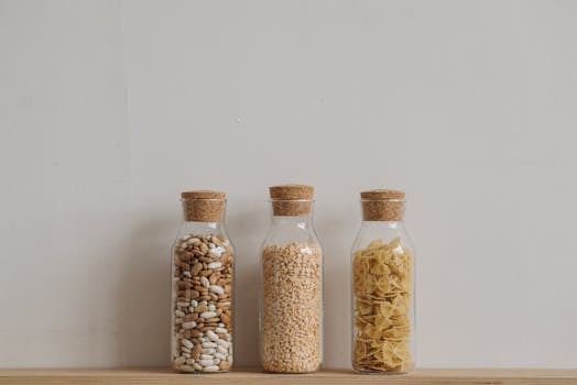 Three glass jars with cork lids holding beans, grains, and pasta on a wooden surface against a neutral backdrop.